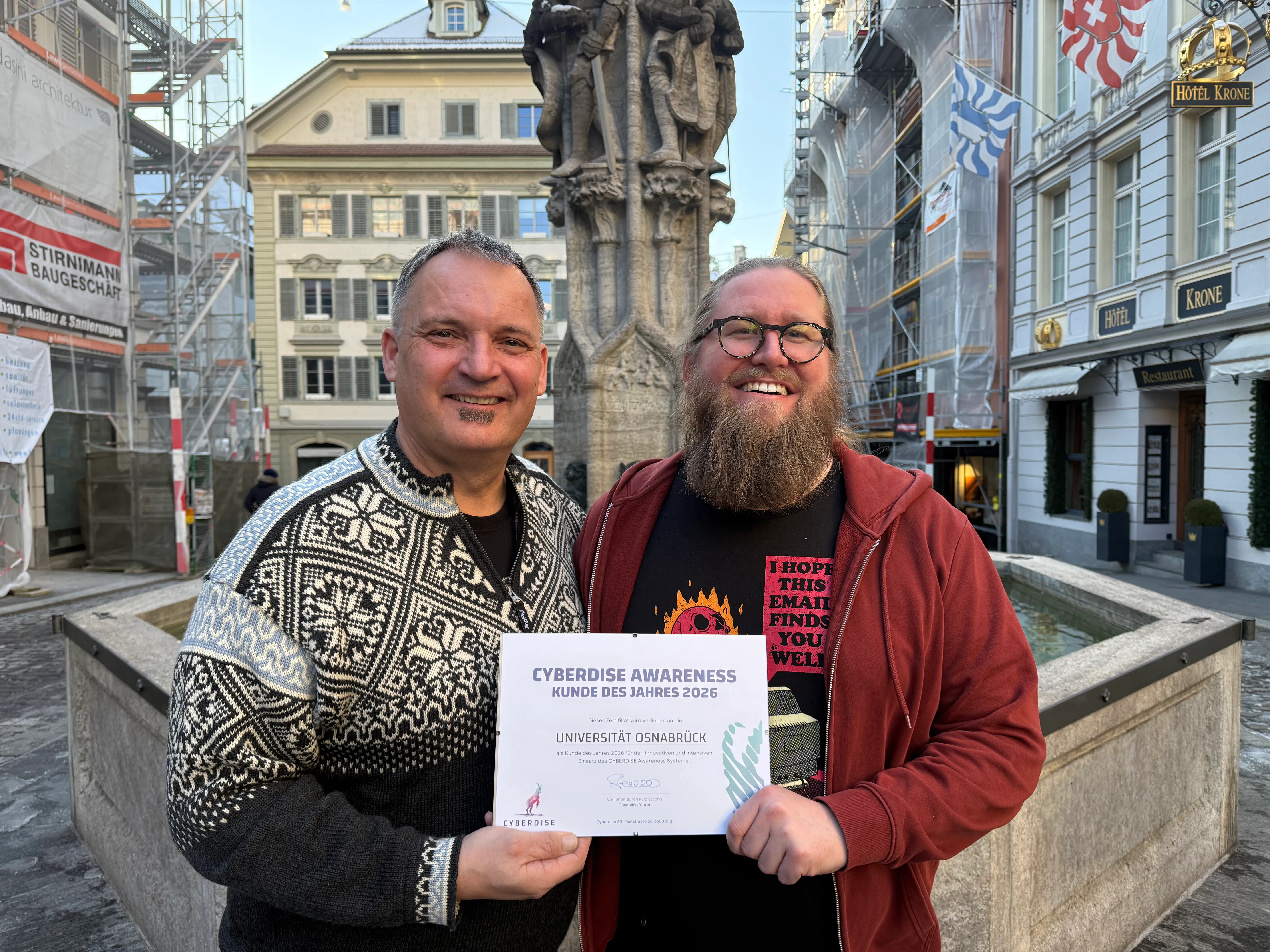 Zwei Männer an einem Brunnen in der Altstadt mit dem Zertifikat «Cyberdise Awareness Kunde des Jahres 2026» für die Universität Osnabrück
